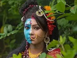 Portraitfoto einer reichlich geschmückten Person, die in die Kamera blickt. Sie trägt Make-Up, das ihr Gesicht optisch in zwei Teile teilt. Die rechte Gesichtshälfte ist blau bemalt.