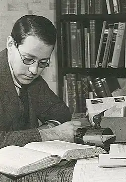 Das Bild zeigt ein Schwarz-Weiß-Foto von Øyvind Anker (1935) an einem Schreibtisch sitzend. Der Schreibtisch ist mit Büchern, Papieren und einem Schreibgerät, wahrscheinlich einer Schreibmaschine, bedeckt. Im Hintergrund befindet sich ein großes Bücherregal, das mit einer Vielzahl von Büchern gefüllt ist. Die Szene wirkt wie ein Arbeits- oder Studienbereich, der eine konzentrierte Atmosphäre ausstrahlt.