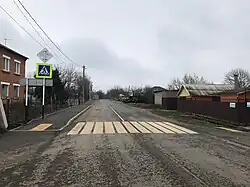 Nichtbefahrene asphaltierte Straße mit Zebrastreifen und mit beidseitiger Bebauung im Panoramablick auf den Himmel nebst Begrünung am Horizont in Farbe.