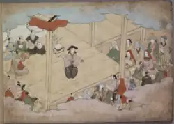 Okuni steht auf einer Bühne, vor der mehrere Zuschauer sitzen. Sie trägt einen schwarzen Kegelhut, ein beigenes Oberteil und eine weite, schwarze Hose mit weißem Pflanzenmuster. In der linken Hand hält sie einen Gong, in der rechten einen schwarzen Gongschlägel. Hinter ihr sitzen mehrere als Priester verkleidete Schauspieler. Sie tragen einen Chonmage, kahl rasiertes Haupthaar plus Pferdeschwanz. Die Priester tragen verschiedenfarbige Gewänder, einer von ihnen schlägt eine kleine Trommel.