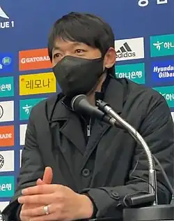 Sitzender Sportler ab Brustkorb mit Mundmaske vor einer Tafel mit Sponsorenbildern.