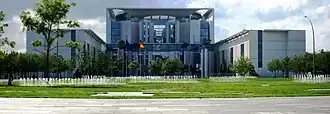 Frontale Farbfotografie vom Bundeskanzleramt mit Rasen und vielen kleinen Wasserfontänen. Vor dem Eingang stehen eine Europaflagge und eine Deutschlandflagge. Das Hauptgebäude besteht aus viel Glas und einigen Betonstützen und wird von zwei quaderförmigen Seitenflügeln flankiert.