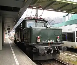 Bild der in grüner Farbe lackierten Museumslok 1020.42‑6, die im Bregenzer Hauptbahnhof auf die Weiterfahrt wartet. Im Hintergrund ist der Nahverkehrstriebwagen 4024 der ÖBB zu sehen.