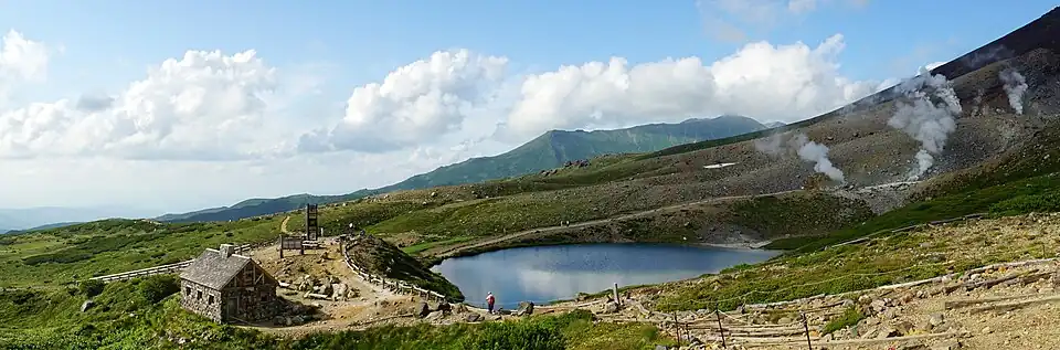 Eine Panorama-Fotografie zeigt eine imposante Landschaft im Gebirge. In der Mitte ist ein kleiner See und links davon ein Steinhaus zu sehen. Der Himmel ist strahlend blau.