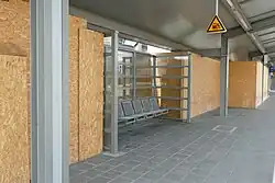 Fotografie eines Bahnsteigs mit einer metallenen Sitzbank. Hinter der Sitzbank sieht man eine Pressspanwand, die sich durch das gesamte Bild zieht.