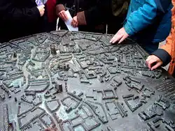 Blick von schräg oben auf ein bronzenes Relief, das modellhaft die Altstadt von Münster zeigt. Es ist in der Fußgängerzone der Münsteraner Altstadt etwa in Hüfthöhe horizontal auf einen Sockel montiert. Von den dicht dahinter stehenden Passanten sieht man nur die Hüften, einige betasten mit ihren Händen die Details des Modells. Dieses zeigt leicht stilisiert Straßen, Plätze, Gebäude und Baumgruppen; die Häuser sind etwa 2&nbsp;cm hoch. Der Dom, die Lamberti- und die Liebfrauenkirche ragen mit mehreren Zentimetern Höhe deutlich heraus. Die Straßen sind in Brailleschrift mit ihren Namen gekennzeichnet. Am rechten Bildrand erkennt man Teile einer Widmung; dieser mehrzeilige Text ist sowohl in Brailleschrift als auch in erhabener Normalschrift ausgeführt.