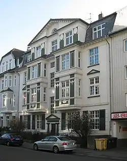 Wohngebäude mit einer aufwendigen Fassade. Das Gebäude hat mehrere Etagen und ist in den Farben Weiß und Grün gestaltet. Die Architektur weist dekorative Elemente auf, darunter Pilaster, Gesimse und Fensterrahmen mit filigranen Designs. Es gibt mehrere Fenster auf jeder Etage, einige davon mit Fensterläden. Der Eingangsbereich wird von einem kleinen Portikus mit einem dreieckigen Giebel betont.Vor dem Gebäude stehen zwei geparkte Autos, und es sind Büsche sichtbar, die möglicherweise Teil eines Gartens sind. Auf der rechten Seite des Gebäudes befindet sich ein Schild mit der Aufschrift „LAGER-VERK“ sowie der Zahl „55“.