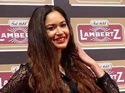 Lovelyn Enebechi bei der Lambertz Monday Night, im Februar 2016. Lovelyn Enebechi mit langen, welligen braunen Haaren, die eine schwarze Spitzenbluse trägt. Sie posiert mit einer Hand in den Haaren. Der Hintergrund ist ein wiederholendes Muster des Lambertz-Logos. Das Logo enthält die Aufschrift „Seit 1688“ und „Lambertz“ in weißen Buchstaben vor einem roten und goldenen Hintergrund.