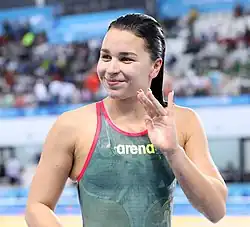 Késely trägt einen grünen Badeanzug mit rotem Rand. Sie winkt nach einem Sieg ins Publikum.