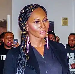 Porträtfoto von Dominique Jackson. Sie trägt einen marineblauen Blazer, eine dunkelblaue Bluse, aufgemalte Augenbrauen, rosa Lidschatten und Lipgloss sowie an beiden Ohren mehrteiligen Schmuck bestehend aus Ohrsteckern, Kugeln und Ohrringen. Ihre schwarzen Haare trägt sie als Braids, also geflochtene Zöpfchen mit goldenen Haarringen. Im Hintergrund stehen einige afroamerikanische Männer vor einer weißen Wand, von denen einige Jackson betrachten.