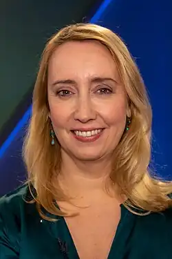 Melanie Amann, Porträt hat blondes schulterlanges Haar. Sie trägt ein dunkelgrünes ärmelloses Oberteil mit V-Ausschnitt, dazu trägt sie kleine, dezent wirkende Ohrringe mit türkisfarbenen Steinen. Der Hintergrund ist unscharf und zeigt blaue und grüne Farbtöne. Das Foto wirkt professionell und wurde bei der Sendung Maischberger 2019 aufgenommen.