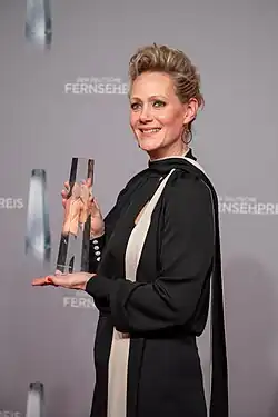 Anna Schudt beim Deutschen Fernsehpreis 2019 in der Düsseldorfer Rheinterrasse, die eine elegante Trophäe, den Deutschen Fernsehpreis, in den Händen hält. Die Trophäe ist rechteckig und aus transparentem Material wie Glas oder Kristall gefertigt, mit klaren Kanten und einer länglichen, prismatischen Form. Anna Schudt trägt ein schickes schwarzes Kleid, das durch einen weißen diagonalen Streifen verziert ist, der von der Schulter bis zur Taille verläuft. Der Hintergrund ist grau und mit dem Schriftzug „DER DEUTSCHE FERNSEHPREIS“ versehen. Anna Schudts Haltung strahlt Stolz und Freude aus, was durch das Präsentieren der Trophäe verstärkt wird. Es ist eine feierliche und besondere Momentaufnahme.