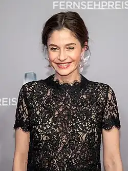 Anne Werner lächelt in einem eleganten schwarzen Spitzenkleid mit kurzen Ärmeln und einem hohen Kragen beim Deutschen Fernsehpreis 2019 in Düsseldorf. Im Hintergrund ist ein grauer Bereich zu sehen, auf dem teilweise das Wort "FERNSEHPREIS" steht.