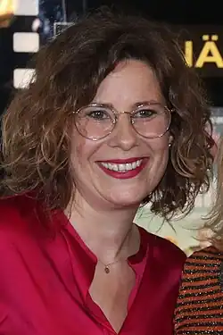 Nicole Kersten lächelt freundlich, mit schulterlangem, lockigem Haar, die eine rote Bluse trägt. Sie hat eine kleine, runde Anhänger-Halskette um den Hals. Im Hintergrund ist ein schwarzes Schild mit gelber Schrift sichtbar, auf dem „Ä“ geschrieben steht. Rechts im Bild ist eine weitere Person teilweise zu sehen, die ein gestreiftes, glitzerndes Oberteil trägt.Das Foto entstand bei der Premiere des Fernsehfilmes „Klassentreffen“ 2019.