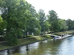 Zu sehen ist der Kanal vorn, ein grüner Uferstreifen und dahinter ein von Bäumen gesäumter Weg. Das Bootshaus ist weitgehend von den Bäumen verdeckt, während der Steg gut erkennbar ist.