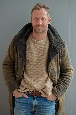 Maximilian Brückner trägt eine braune Jacke mit einem dunklen, pelzartigen Kragen. Darunter trägt er einen beigen Strickpullover, der einen gemütlichen Eindruck vermittelt. Maximilian Brückner hat die Hände in den Taschen einer blauen Jeans gesteckt, die mit einem braunen Gürtel getragen wird. Der Hintergrund des Bildes ist schlicht und einfarbig grau. Er lächelt leicht.