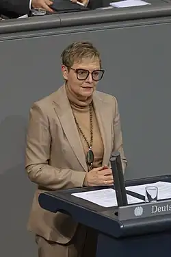 Sabine Dittmar steht an einem Pult und trägt kurzes blondes Haar. Ihr Anzug ist beigefarben und sie trägt eine Brille.