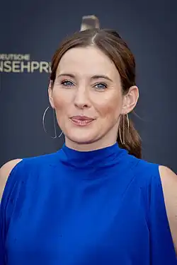 Jeannine Michaelsen auf dem roten Teppich des Deutschen Fernsehpreises 2022, trägt ein blaues Oberteil und hat eine lächelnde Miene. Sie steht vor einem Hintergrund mit teilweise sichtbarem Text "DEUTSCHER FERNSEHPREIS". Sie trägt ein ärmelloses, leuchtend blaues Oberteil mit hohem Halsausschnitt. Die Haare von ihr sind nach hinten gestylt, und große Kreolen sind sichtbar. Der Hintergrund scheint dunkel zu sein, möglicherweise schwarz oder dunkelblau, mit dem Logo des Deutschen Fernsehpreises