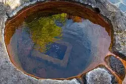 Runder, steinumfasster Brunnen mit rostroten Ausfällungen und aufsteigenden Bläschen. Unter Wasser ist eine weitere, rechteckige Einfassung erkennbar