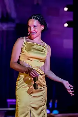 Gina Haller in einem goldenen Kleid, lächelnd auf der Bühne der Nibelungen Festspiele 2022 in Worms, mit einer Sonnenbrille in der Hand. Gina Haller trägt ein elegantes, bodenlanges Kleid in einer dunklen Farbe, das durch seinen schlichten und klassischen Schnitt besticht. Ihr Haar ist stilvoll hochgesteckt. Sie steht vor einem Hintergrund, der die Atmosphäre der Festspiele widerspiegelt, möglicherweise mit einem Banner oder einer Kulisse, die auf das Thema der Nibelungen hinweist. Ihr Ausdruck ist freundlich und selbstbewusst, passend zu einem kulturellen Ereignis von großer Bedeutung.