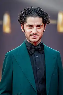 Mann im grünen Sakko, schwarze Hemd, schwarze Fliege, vor Sponsorenwand