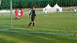 Von hinten durch ein Fußballtor fotografiert, sieht man die Torhüterin der koreanischen Frauenmannschaft im schwarzen Trikot von hinten. Auf dem Spielfeld mehrere koreanische Spielerinnen in Rot und Spielerinnen der Elfenbeinküste in weißem Shirt und grüner kurzer Hose, im Hintergrund weiße Zelte.