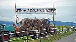Ein Bild eines Traktors mit einem Anhänger voller Heuballen. In den Heuballen stecken Schuhe und Fahrradteile, als ob sie mit hoher Geschwindigkeit hineingefahren wären. Darüber ein Transparent mit der Aufschrift «Wenn das UCI schlecht informiert».