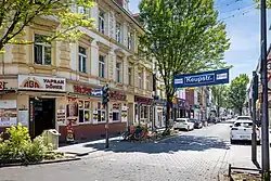 Das Foto zeigt eine Straße mit mehrstöckigen Häusern auf beiden Seiten, aufgenommen bei Tageslicht unter blauem Himmel. Die Gebäude links und rechts haben helle oder pastellfarbene Fassaden, teils mit Schildern und Markisen. Im Vordergrund steht ein blaues Straßenschild mit der Aufschrift „Keupstr.“ über der Straße. Auf beiden Straßenseiten sind zahlreiche Autos parallel geparkt, darunter weiße, silberne und dunkle Fahrzeuge. Links im Bild befindet sich ein Döner-Imbiss mit der Aufschrift „Yaprak Döner“, daneben weitere Geschäfte mit auffälliger Beschriftung, unter anderem ein Laden mit rotem Leuchtschild „Chef“. Einige Fahrräder stehen am Rand. Bäume mit grünem Laub stehen beidseitig der Straße, ihre Kronen reichen teilweise über die Fahrbahn. Stromleitungen und Lichterketten spannen sich über die Straße hinweg. Die Perspektive zeigt die Straße in Tiefe und verjüngt sich leicht zum Horizont hin.