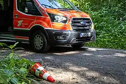 Ein liegender Leitkegel auf einem Waldweg, dahinter ist ein Personentransporter der Feuerwehr zu sehen.
