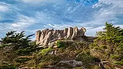 Große, markante Felsformation auf einem bewachsenen Hügel, umgeben von niedrigem Gestrüpp und schräg gewachsenen Bäumen. Die Felsen wirken gezackt und säulenartig, fast wie eine steinerne Krone. Darüber ein teils bewölkter Himmel mit blauen und weißen Farbtönen.