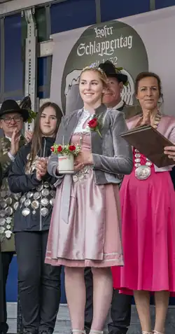 Foto einer Frau auf der Bühne. Im Hintergrund stehen weitere Personen. Junge Frau hat Krug mit Aufschrift „Schlappenbier“ in der Hand. Guckt lächelnd nach links.