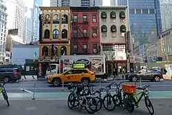 Alleinstehendes mehrstöckiges Haus aus braunen Ziegeln mit rechts einem Reklameschild für einen „Celtic Pub“ und link einem für eine Trattoria. Vor dem Haus steht ein Lastwagen und ein gelbes Taxi, dahinter höhere Hochhäuser.