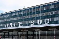 Das Foto zeigt einen Teil der Fassade eines vier- oder fünfgeschossigen Gebäudes mit Flachdach. Die Vorhangfassade ist dominiert von Glas und Metall. Die oberen drei Stockwerke enthalten offenbar Büroräume, wie man durch die Fensterbänder erkennen kann. Auf einem Vordach steht in großen Lettern „Orly Sud“, was das Gebäude als Flughafenterminal erkennbar macht.