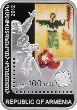 Eine hochkant stehende rechteckige Münze. Darauf sind eine Kamera, ein Bild mit einem grün gekleideten Mann, ein aufgeschnittener Granatapfel und ein Wappen abgebildet. Dazu der „Republic of Armenia“ und „DRAM&nbsp;100“
