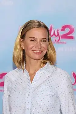 Nadeshda Brennicke bei der Premiere von Wendy 2 im Februar 2018. mit schulterlangem blondem Haar, die ein hellblaues Hemd mit kleinen roten und blauen Punkten trägt. Der Hintergrund des Bildes ist hellblau, und darauf sind Textfragmente des Schriftzuges Wendy zu sehen, darunter eine große rosa Zahl „2“ und der weiße Schriftzug „IM KINO“.