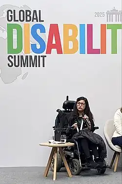 Abia Akram sitzt in ihrem schwarzen elektronischen Rollstuhl auf einer Bühne. Sie hat dunkles langes, gewelltes Haar und trägt eine Brille. Im Hintergrund ist eine weiße Wand, auf der in bunten Buchstaben „Global Disability Summit 2025“ steht.
