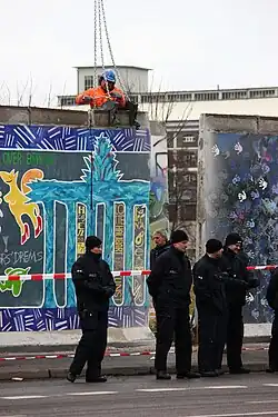 Frontale Farbfotografie von der Entfernung der Berliner Mauer mit schwarz gekleideten Polizisten und einem Absperrband im Vordergrund. In der Mitte ist ein Mauerstück nach hinten versetzt worden und ein Bauarbeiter befestigt eine Hebevorrichtung mit Ketten an dem linken Mauerstück an.