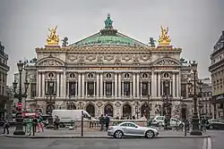 Frontale Farbfotografie eines zweigeschossigen Gebäudes im Neobarock-Stil. Im Erdgeschoss sind Rundbögen und im Obergeschoss sind zwischen den Fenstern Säulen, über denen „Académie nationale de musique“ steht. An den Dachecken stehen zwei goldene Figuren und das Gesims ist Gold. In der Mitte ist eine grüne Kuppel mit Figuren, die eine goldene Krone hochhalten.