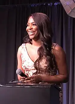 Alexandra Grey steht lächelnd an einem schwarzen Rednerpult und hält ein schwarzes Mikrofon in der rechten Hand. Sie trägt ein weißes, ärmelloses Kleid mit braunen Vogelaufdrücken. Ihre braunen Haare trägt sie auf der linken Seite bis zur Brust reichend und gewellt, auf der rechten Seite nach hinten gekämmt. Hinter ihr ist ein dunkellila Theatervorhang zu sehen.