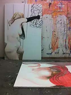 "Das Gemälde zeigt das Atelier einer Künstlerin. Eine unbekleidete Frau mit blondem Haar trägt schwarze Handschuhe und schlägt mit der rechten Faust auf ein Gemälde an der Wand. Dahinter mit Farbe bespritzte Leinwände. Auf dem Boden liegt ein Gemälde eines weiblichen nackten Oberkörpers, der nach vorn gebeugt ist, das Gesicht ist mit roten Haaren oder einem roten Tuch bedeckt."