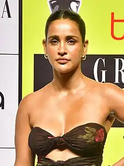 Aisha Sharma mit langen, lockigen Haaren in einem blauen, tief ausgeschnittenen Kleid. Sie trägt ein blaues Kleid mit einem Neckholder-Ausschnitt. Der Ausschnitt hat ausgeschnittene Details im Brustbereich. Im Hintergrund sind große, teilweise sichtbare weiße und rote GQ -Buchstaben auf einem dunklen Hintergrund zu erkennen.