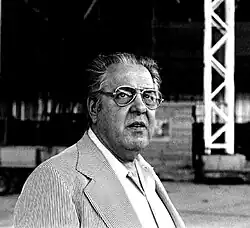 Albert R. Broccoli ist auf einer quadratischen schwarz-weiß Fotografie abgebildet. Er trägt eine Brille und hat wenig Haar. Er trägt einen hellen Anzug.