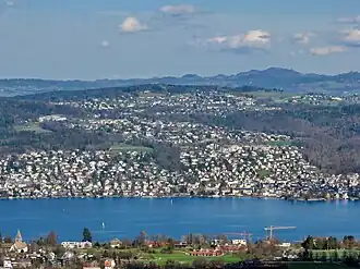 Blick von Westen über den Zürichsee auf die besiedelte Westflanke; von unten: Küsnacht (Dorfzentrum am See), Itschnach (Ortsteil von Küsnacht), Zumikon (Bildmitte, mit höchstem Punkt Chapf) und Forch (rechter Bildrand)