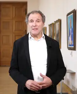 Das Foto zeigt Alexander Hold frontal mit schwarzem Jackett und weißem Hemd. Seine Hände berühren sich und er schaut konzentriert mit offenem Mund in die Kamera.