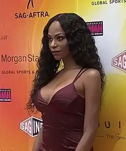 Grey steht vor einer orange-gelben Wand mit den schwarzen Aufschriften SAG-AFTRA, Morgan Stanley und Global Sports sowie der rot-weißen Aufschrift SAG. Sie trägt ein dunkelrotes Kleid mit dünnen Trägern und ihre gewellten schwarzen Haare auf der linken Seite nach hinten gekämmt, auf der rechten Seite bis zur Brust gehend.