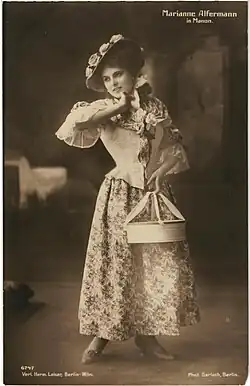 Frau im mittleren Alter in einem zeitgenössische knöchellangen gemusterten Kleid mit Hut und einer Schachtel in der rechten Hand auf einer Bühne stehend von vorne in schwarzweiß.