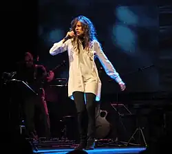 Alice mit langen, lockigen Haaren trägt ein weißes Hemd und schwarze Leggings. Sie steht auf einer Bühne und hält ein Mikrofon, während sie performt. Im Hintergrund ist die Bühne in Dunkelheit getaucht, wobei blaue Lichteffekte für eine stimmungsvolle Beleuchtung sorgen. Links im Bild ist eine weitere Person sichtbar, die Gitarre spielt. Auf der Bühne sind außerdem weitere Musikinstrumente zu sehen, darunter eine Gitarre und ein Keyboard. Die Szene vermittelt die Energie und lebendig Atmosphäre des Live-Konzerts.