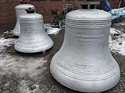 Drei silbern angestrichene, abgehängte Glocken, ringsherum liegt etwas Schnee vor einer Backsteinwand. Auf der vordersten Glocke steht: Kriegsersatz, 1918. Die Liebe höret nimmer auf. 1. Kor. 18,8.