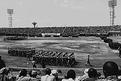 Ein unüberdachtes, auf den Rängen dicht gefülltes Stadion. Auf der Rasenfläche in der Mitte stehen Gruppen von Soldaten in rechteckigen Formationen. Weitere laufen im Gleichschritt auf der Leichtathletikbahn um diese. An den Banden des Stadions steht "Isoko Yo Kwigira", "Kwibohora 20" und "Together We Prosper".