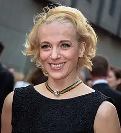 Amanda Abbington mit blonden, lockigen Haaren und einem schwarzen Kleid, trägt eine bunte Halskette, lächelt freundlich. Sie trägt ein elegantes schwarzes Kleid mit kunstvollen Stickmustern. Das Kleid hat ein ärmelloses Design und wirkt stilvoll und raffiniert. Sie hat blondes Haar, das in lockeren Wellen gestylt ist. Um den Hals trägt sie eine auffällige, bunte Halskette mit einem Regenbogenmuster. Im Zentrum der Halskette befindet sich ein dekoratives Element, das wie ein kleines Schmuckstück oder eine Brosche aussieht.Der Hintergrund ist unscharf, es wurde 2015 Laurence Olivier Awards Man aufgenommen. Man erkennt einige weitere Personen in der Umgebung, was darauf hindeutet, dass die Szene vor der Veranstaltung aufgenommen wurde.